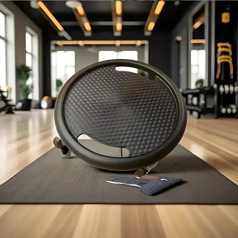 Close-up of a black abdominal gliding disc with a textured surface placed upright on a yoga mat in a modern gym, with natural light and fitness equipment in the background.