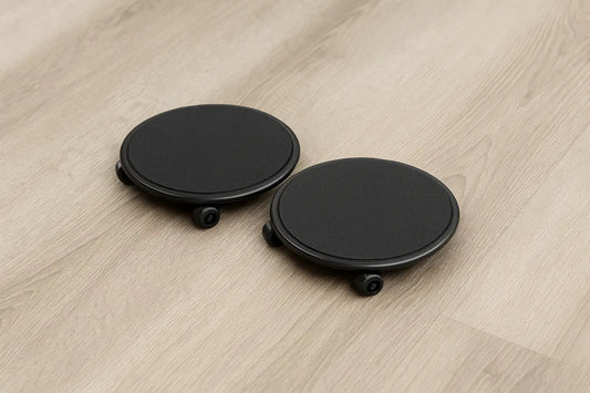 A pair of black abdominal gliding discs with wheels placed on a light wood floor, shown in a close-up view with soft natural lighting highlighting their round shape and smooth surface.